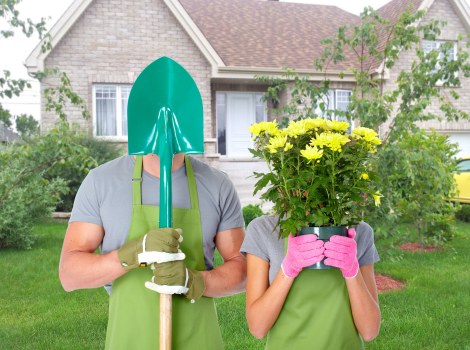A gardener and a woman are standing outdoors in a residential garden, both wearing green gardening aprons and gloves. The man is holding a bright green shovel with a purple handle, with the shovel's blade covering his face. The woman is holding a vibrant yellow flowering plant in a pink container, obscuring her face. Behind them, there is a well-maintained lawn with lush green grass, bordered by a variety of shrubs and small trees. In the background, a house with a beige brick exterior and white window frames is visible, along with a garden bed edged with soil and some mulch. The scene appears to be bright and clear, suggesting a sunny day, suitable for gardening activities. The overall environment reflects a typical suburban garden, ideal for lawn care and planting services offered by Lawn Mowing Lee in the Lee area, with the setting emphasizing outdoor maintenance and horticultural work.